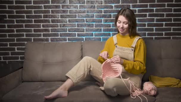 Attractive young caucasian woman in casual clothes sitting on a sofa indoors knitting wool — Vídeo de stock