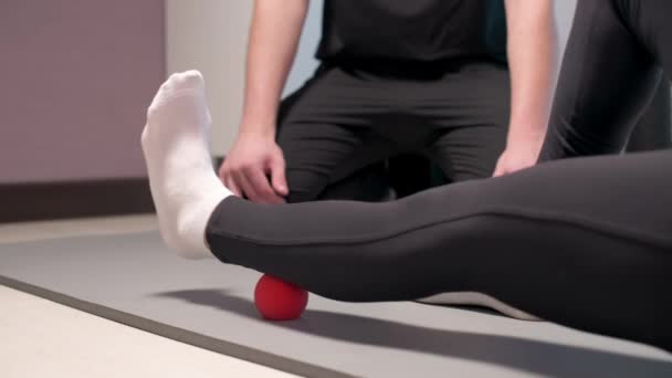 Close-up Joven masajista masculino enseña cliente mujer caucásica paciente miofascial auto-masaje de piernas y caderas utilizando bola de masaje en el interior. Liberación miofascial — Vídeos de Stock