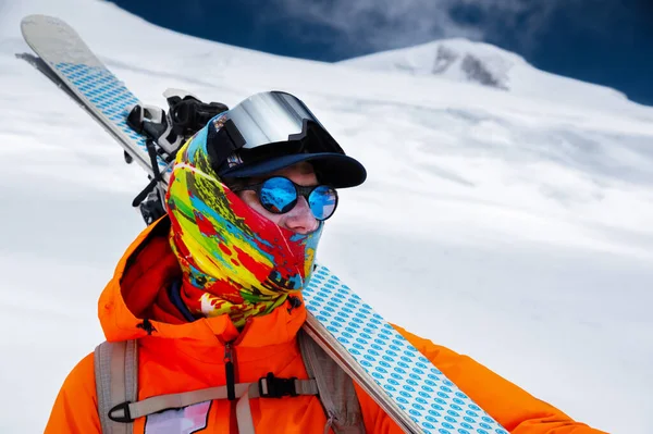 Mountain portrait of a professional freeride skier in orange clothing with ski poles and skis on his shoulders. Stands high in the mountains on a snowy slope — Stock Photo, Image