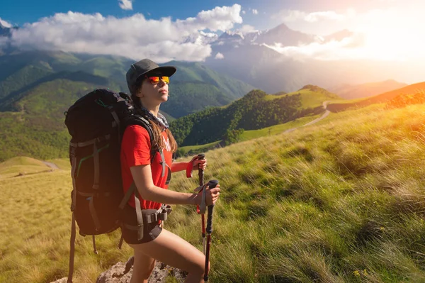 A tourist girl with a backpack admires the sunset from the mountainside. Traveler in the background of mountains and clouds at sunset. Woman tourist in sunglasses, cap and with a backpack — Foto Stock