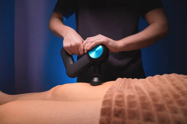Zbliżenie profesjonalnego masażysty męskiego stymuluje mięśnie nóg męskiego pacjenta w ciemnym gabinecie spa do masażu. Perkusja mechaniczny wpływ na przeciążone mięśnie — Zdjęcie stockowe