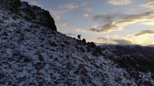 İki turistin hava manzaralı siluetleri, erkek ve kadın dağların tepesinde, batan güneşin ve karla kaplı dağların arka planında dikiliyorlar. — Stok video