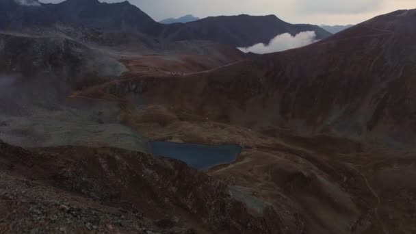 Veduta aerea della cima di una montagna rocciosa con catene montuose e un lago con nuvole basse. Tempo di sera scalando le montagne del Caucaso — Video Stock