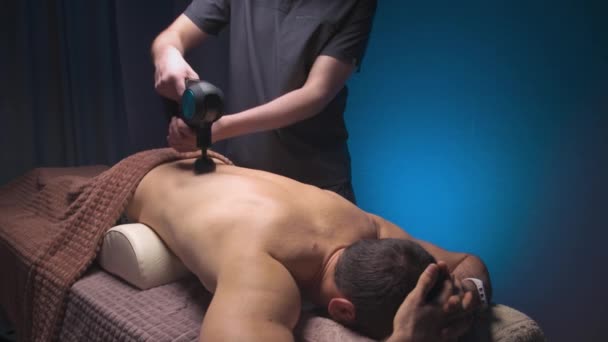 Bir erkek ve bir atlet için perküsyon masaj aleti olan profesyonel masaj, bir kaplıca salonunun karanlık odasında bir masör tarafından yapılır. — Stok video