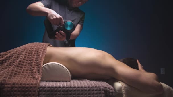 Massage professionnel avec un masseur de percussions pour un homme et un athlète se fait par un masseur dans une pièce sombre d'un salon de spa — Video