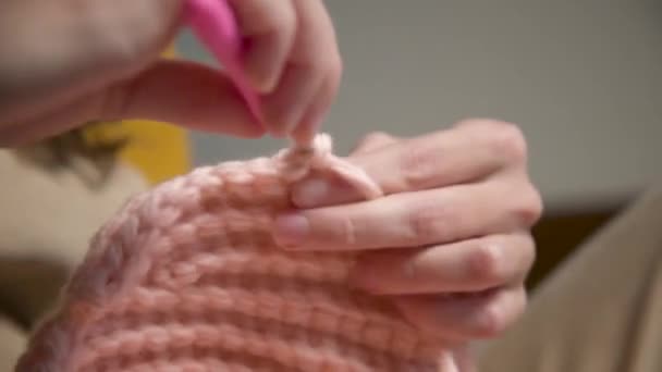 Close-up. Mãos de uma jovem mulher branca atraente em roupas casuais sentado em um sofá dentro de casa tricota um produto de lã por um crochê — Vídeo de Stock