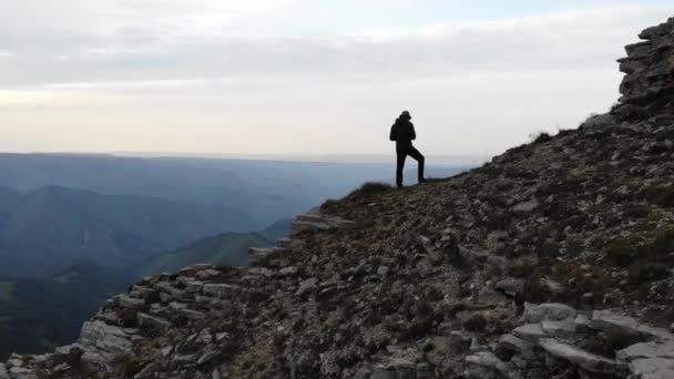 Um homem de roupas de trekking está à beira de um penhasco em frente a um vale da montanha. Controla o voo do drone. Vista aérea — Vídeo de Stock