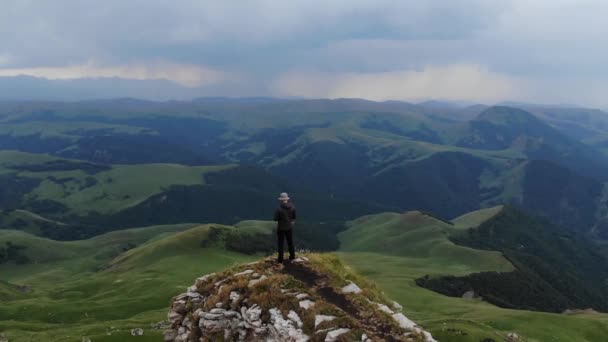 A man in trekking clothes stands on the edge of a cliff in front of a mountain valley. Controls the flight of the drone. Aerial view. Flying around — 비디오