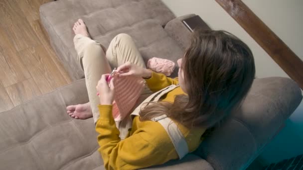 Mujer caucásica joven está tejiendo un producto de lana mientras está sentado en el sofá en casa. Producción casera de prendas de lana — Vídeo de stock