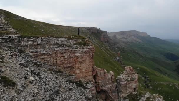 Чоловік у пішохідному одязі стоїть на краю скелі перед гірською долиною. Керує польотом безпілотника. Вид з повітря — стокове відео