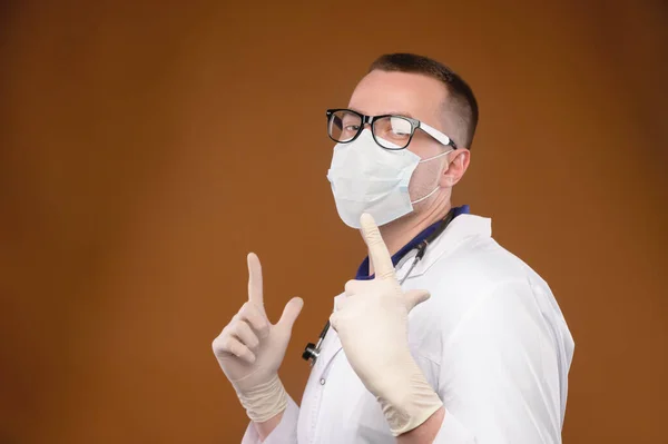 Een vrolijke blanke dokter in een witte jas met bril en een masker danst in de studio met zijn wijsvingers omhoog.. — Stockfoto