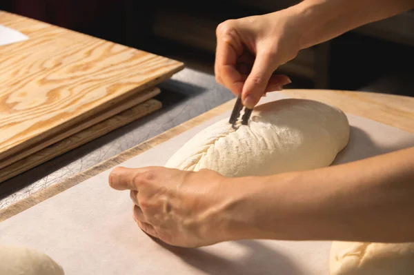 Primer plano de un panadero manos femeninas cortar una hogaza de masa en una hogaza de pan con una hoja antes de hornear en el horno. Producción artesanal de pan artesanal — Foto de Stock