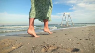 Moda çekimi: kumlu bir plajın dalgalı yüzeyinde yürüyen ince kadın bacaklarının yakın çekimi. Yeşil, pohpohlayıcı elbiseli bir kadın model ufka doğru yürüyor. Yavaş çekimde