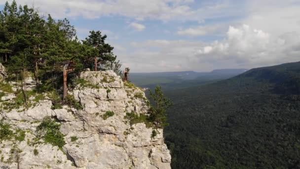 Αεροφωτογραφία ενός νεαρού Καυκάσου με μακριά μαλλιά στέκεται στην άκρη ενός βράχου ψηλά σε ένα βράχο με φόντο μια κοιλάδα στα βουνά καλυμμένη με πράσινο δάσος. Βίντεο ταξιδιού — Αρχείο Βίντεο