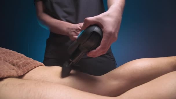 Die Nahaufnahme eines professionellen männlichen Masseurs stimuliert die Beinmuskulatur eines männlichen Patienten in einem dunklen Wellnessraum zur Massage. Percussion mechanische Wirkung auf überbeanspruchte Muskeln — Stockvideo