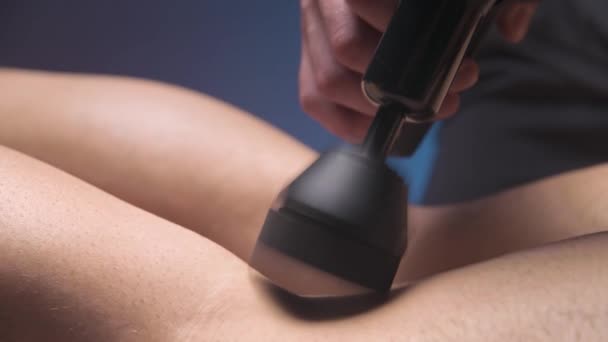 Close-up de um massagista masculino profissional estimula os músculos das pernas de um paciente masculino em uma sala de spa escura para massagem. Efeito mecânico da percussão nos músculos sobrecarregados — Vídeo de Stock