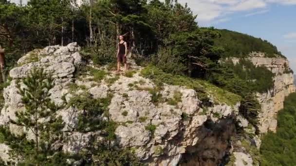 Vue aérienne d'un jeune homme caucasien aux cheveux longs se dresse sur le bord d'une falaise haute sur un rocher sur fond de vallée dans les montagnes couvertes de forêt verte. Vidéo de voyage — Video