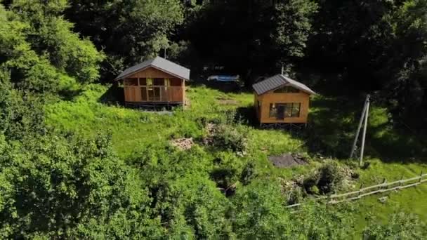 Вид с воздуха два деревянных дома стоят на газоне в окружении зеленого леса. Эко поселение далеко в горах. Современные места для отступления и понижения — стоковое видео