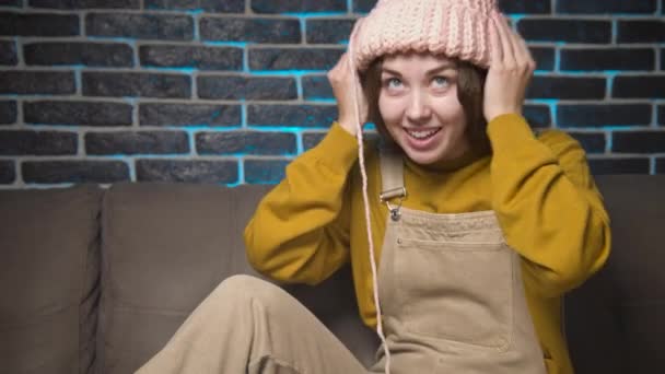 Feliz mujer caucásica atractiva riendo probándose una gorra de punto mientras está sentado en un sofá en una habitación — Vídeo de stock