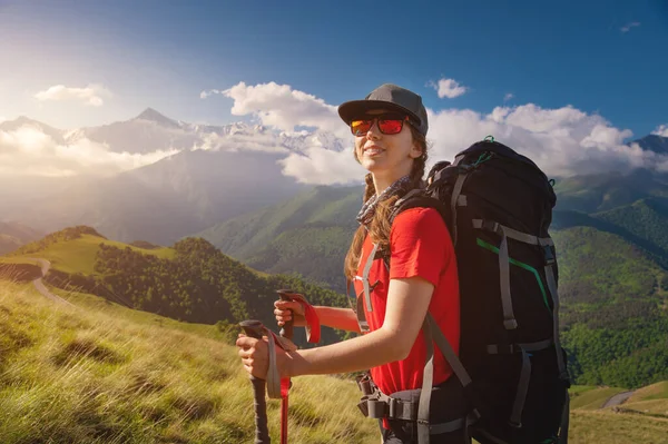 Kaukázusi utazó sapkában és napszemüvegben, hátizsákkal a hátán, és botokkal a hátán, magas hegyekkel és alacsony felhőkkel. Mosolyog és a kamerába néz. — Stock Fotó