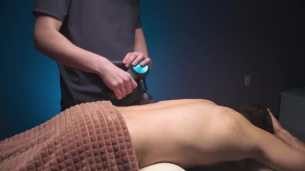 Кавказький професійний масажист робить масаж для нижньої спини і квадратного м'яза спини чоловічого клієнта, використовуючи масажер вібрації. Шокова терапія для регенерації масажу — стокове відео