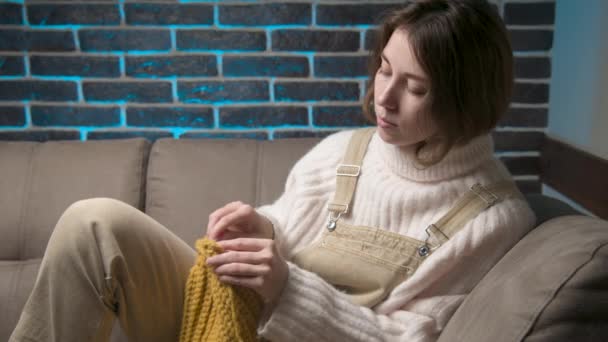 Een jonge blanke aantrekkelijke vrouw in casual huiskleding zit op de bank in de kamer en breit een hoed van wol. Hobby en thuiskleding maken — Stockvideo