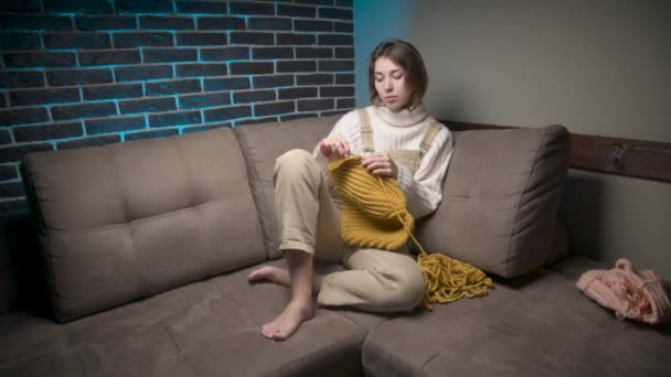 Attractive Caucasian young woman in casual clothes knitting a wool hat while crocheting while sitting on the sofa at home. Handicraft hobbies and home clothing making — Stock Video