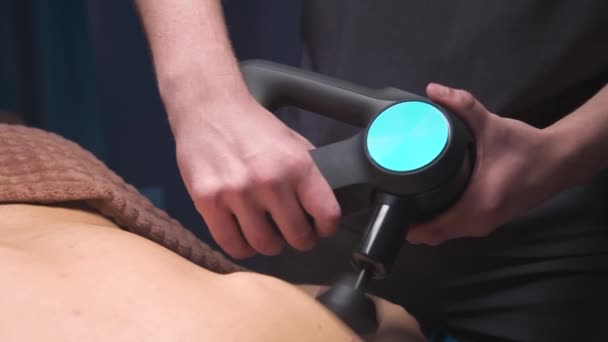 Primo piano massaggiatore di percussioni nelle mani di un massaggiatore fa un massaggio delle spalle e parte superiore della schiena ad un cliente maschio sdraiato — Video Stock