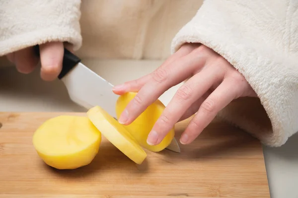 Joven mujer caucásica en un albornoz se para y corta patatas en una tabla de cortar — Foto de Stock