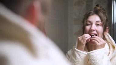 Genç bir kadın, banyo aynasına bakarken üst dişlerini düzeltmek için alt dişlerine düzenleyici bir sabitleyici takıyor. Düzeltmek için alt dişlere hizalayıcı kurulumu