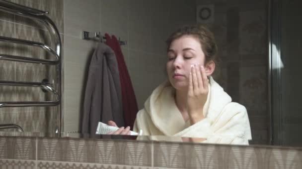 Aantrekkelijke jonge blanke vrouw in de badkamer voor een spiegel brengt crème op haar gezicht. Gezichtsverzorging. Massabewegingen — Stockvideo