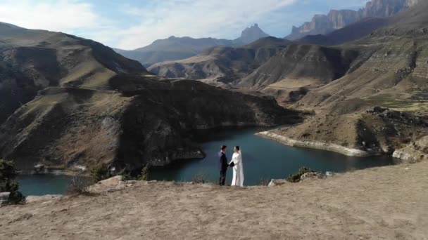 Vista aérea de um casal apaixonado à beira de um penhasco perto da água de um lago de montanha. Uma mulher com um vestido branco incrível e um homem de fato. Pessoas recém-casados em uma caminhada de casamento — Vídeo de Stock