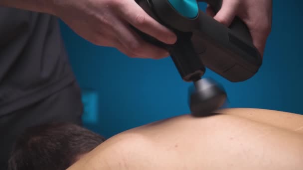 Massagista masculino profissional caucasiano faz um efeito curativo nos músculos da parte superior das costas de um cliente masculino usando um massageador de vibro de percussão. Terapia de choque para regenerar o corpo atlético de massagem. — Vídeo de Stock