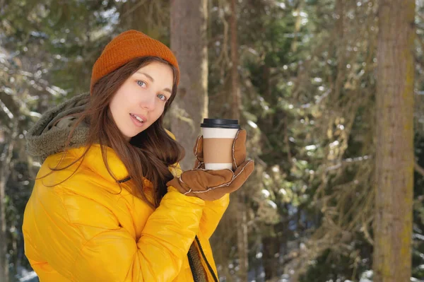 冬服の若い優しい白人女性がカメラと笑顔を見ています。コーヒーを入れた紙コップを鑑賞者に提示します。森の中で冬のホットドリンクを宣伝。ロゴブランクデザイン — ストック写真