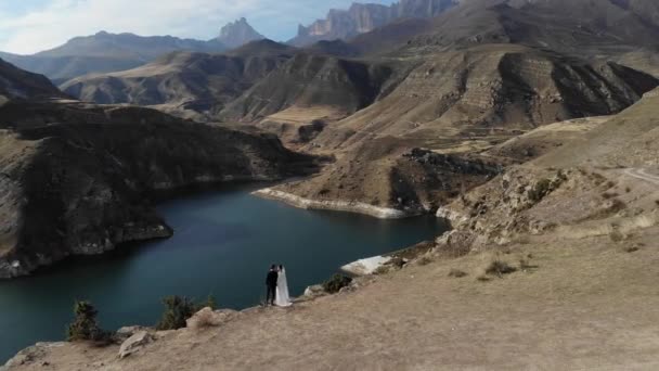 Vista aérea de um casal apaixonado à beira de um penhasco perto da água de um lago de montanha. Uma mulher com um vestido branco incrível e um homem de fato. Pessoas recém-casados em uma caminhada de casamento — Vídeo de Stock