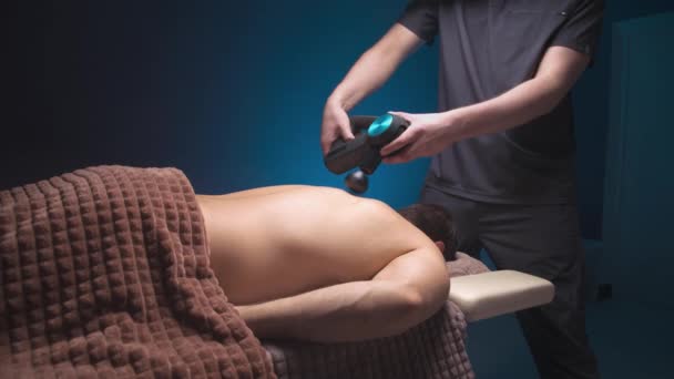 Massageador de percussão nas mãos de um massagista faz uma massagem dos ombros e parte superior das costas para um cliente masculino deitado — Vídeo de Stock