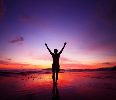 Young man standing on tropical beach with raised hands at sunset