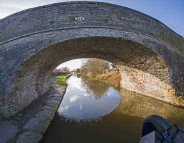Dar kayığın pruvasından İngiliz kırsal manzarasını gösteren eski taş yol köprüsü olan İngiliz su yolu kanalı manzarası.
