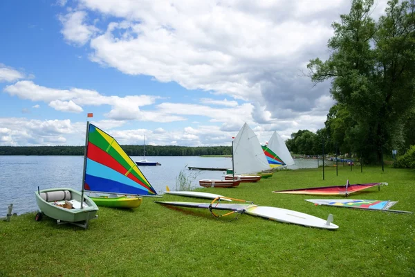 Vela, veleros Optimistas y tablas de Windsurf, extendidas a orillas del lago — Foto de Stock