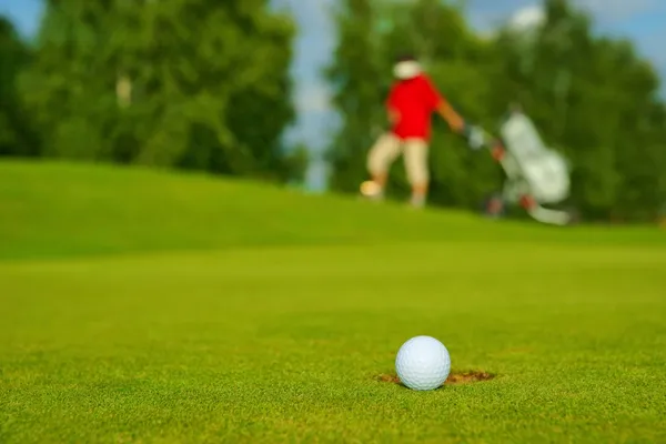 Golf, palla sdraiato sul verde accanto al buco, sullo sfondo va golfista Immagini Stock Royalty Free