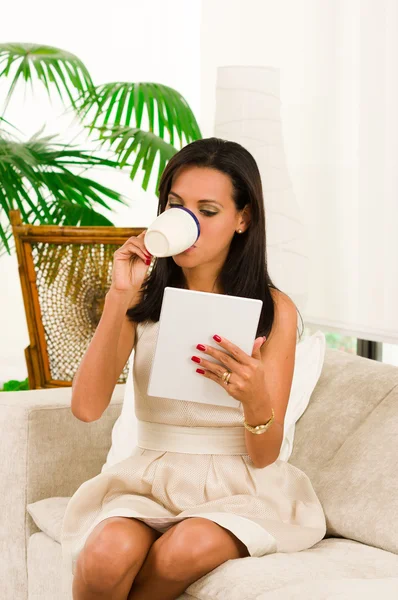 Mooie elegante vrouw zitten in de Bank met behulp van Tablet PC en het drinken van een kopje koffie — Stockfoto