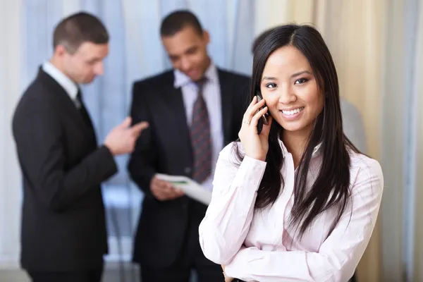 Feliz joven asiática mujer en el teléfono con su equipo de trabajo en el fondo —  Fotos de Stock