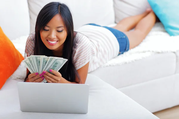 Happy asian woman holding stack of dollars while using her laptop — Stock Photo, Image