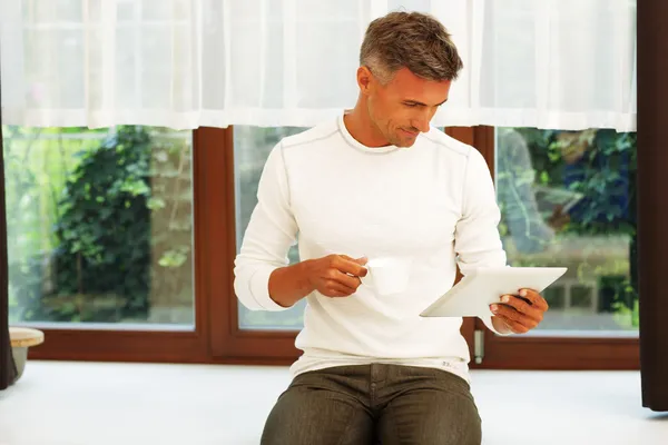 Hombre en casa bebiendo café y leyendo noticias en su tableta electrónica —  Fotos de Stock