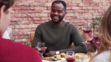 Gülümseyen Afro-Amerikalı bir adam tatilini arkadaşlarıyla kutluyor. Dışarıda, evin yakınındaki terasta şarap bardakları tokuşturuyor.