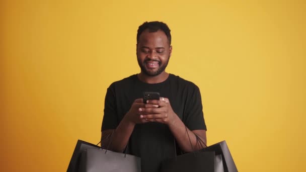 黄色の背景に黒いショッピングバッグで隔離された電話スタンドを使用して笑顔アフリカ系アメリカ人男性 — ストック動画