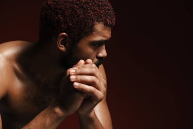 Siyah tişörtsüz sakallı adam kırmızı arka planda yalnız poz veriyor ve yan yana bakıyor.