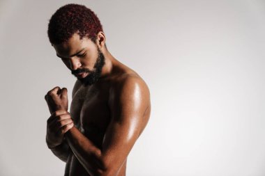 Siyah tişörtsüz sakallı adam beyaz arka planda poz veriyor ve aşağıya bakıyor.