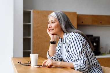 Mutlu, beyaz saçlı, olgun bir kadın kahve içiyor ve cep telefonunu kapalı bir yerde kullanıyor.