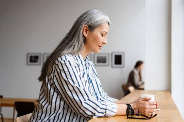 Masada otururken kahve içen beyaz saçlı, olgun bir kadın.